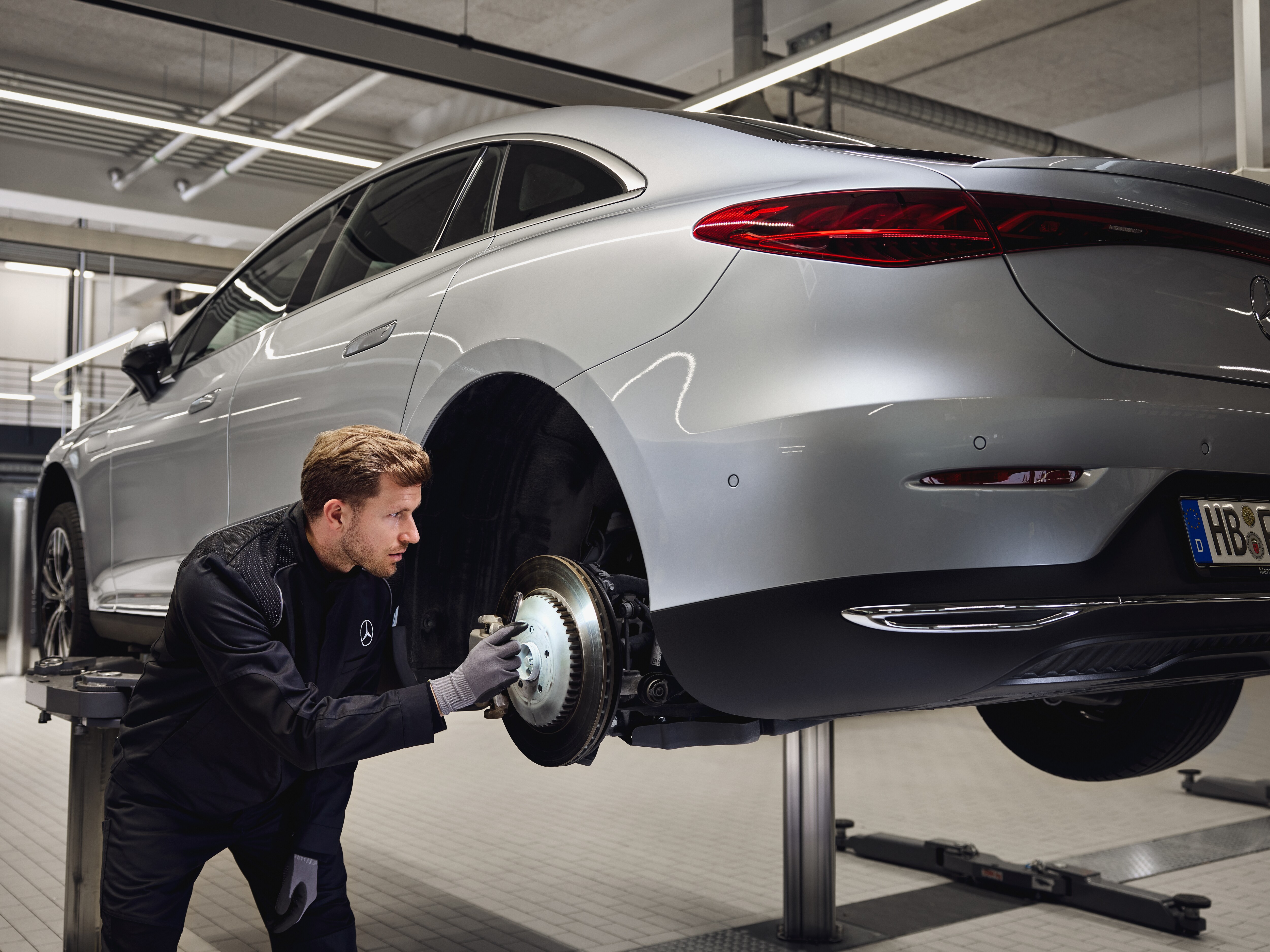 Un véhicule Mercedes-Benz surélevé sur un pont élévateur, avec un technicien en train d'inspecter le système de freinage arrière dans le cadre d'une maintenance avec remplacement de pièces d'origine.