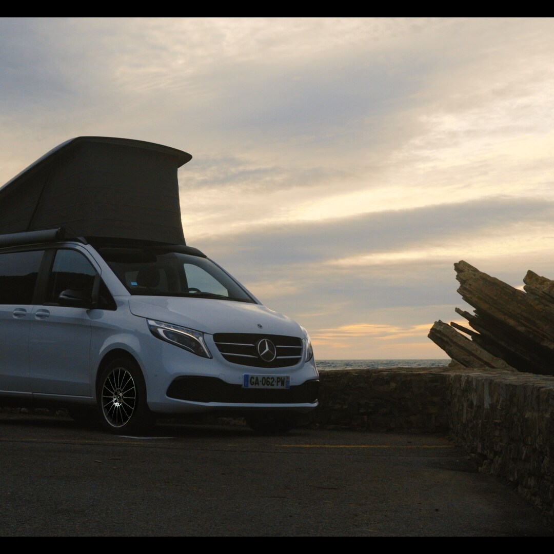 Mercedes-Benz Classe V Marco Polo stationnée en bord de mer avec le toit relevable ouvert au coucher du soleil.