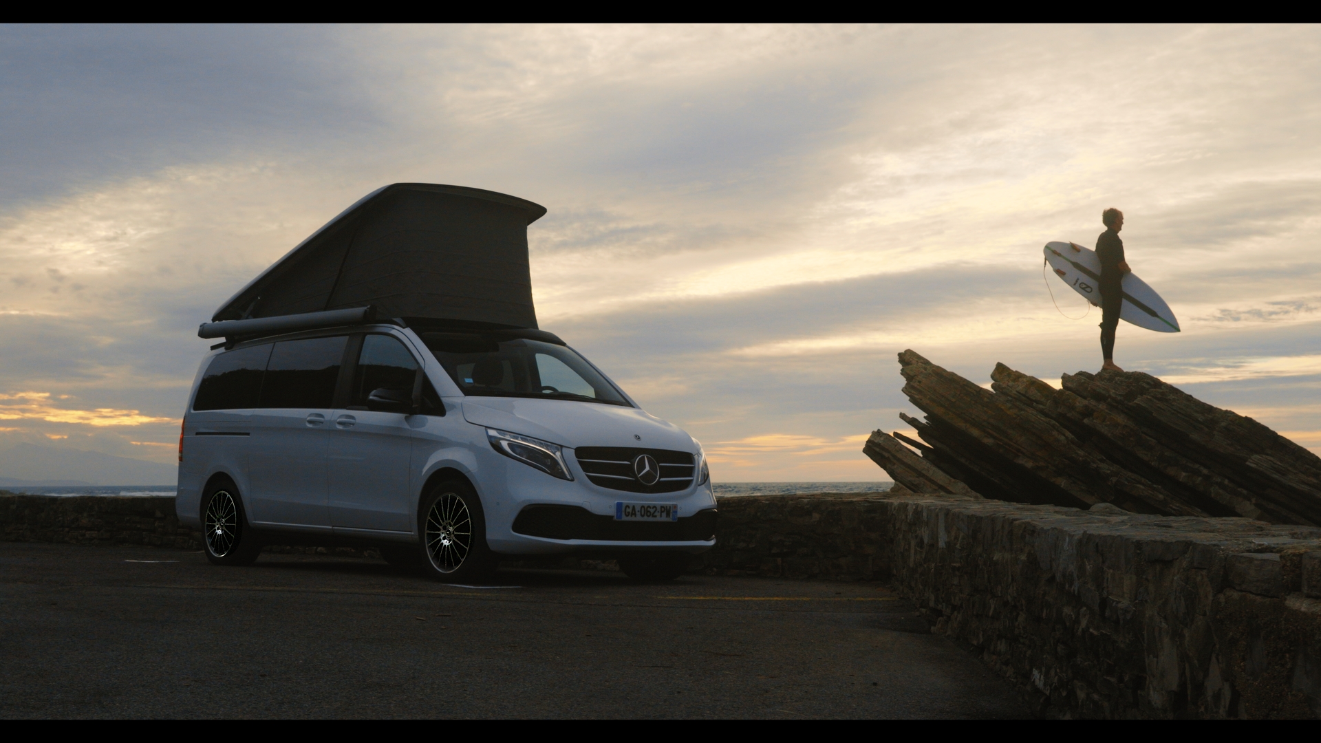 surf Mercedes-Benz Classe V Marco Polo stationnée en bord de mer avec le toit relevable ouvert au coucher du soleil.