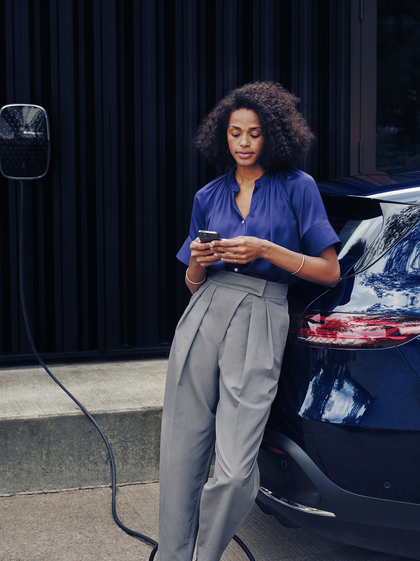 Une femme regarde son téléphone portable à coté d'une Mercedes Benz EQC bleue