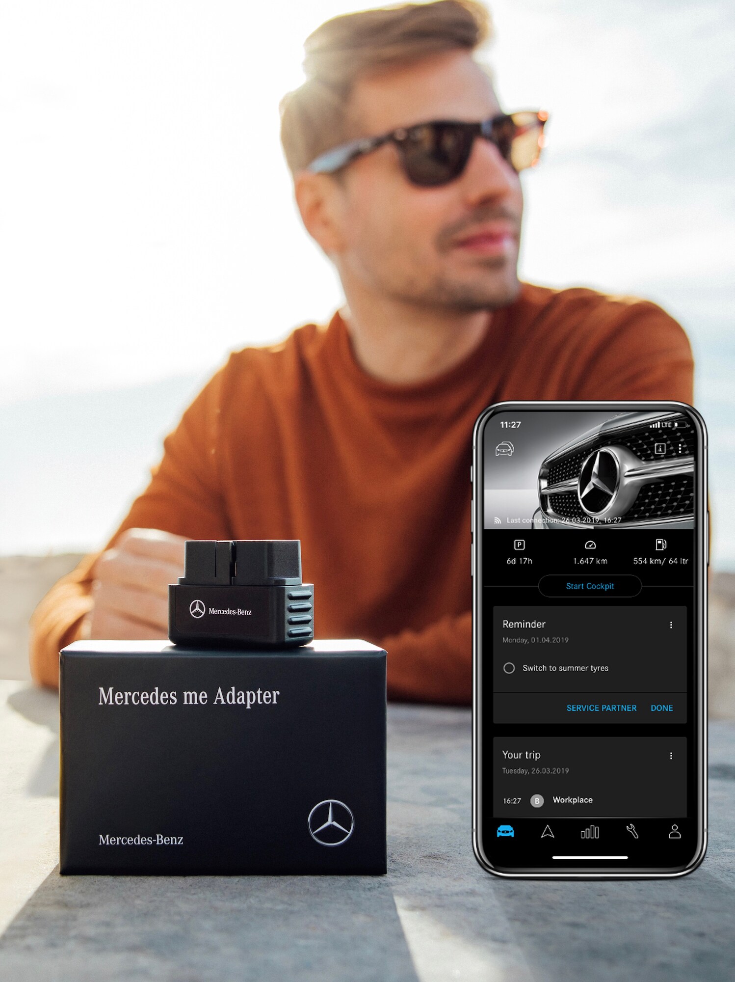 Un homme portant des lunettes de soleil est assis à une table sur laquelle se trouvent, au premier plan, des adaptateurs Mercedes me et un smartphone avec l'application Mercedes me Adapter ouverte.
