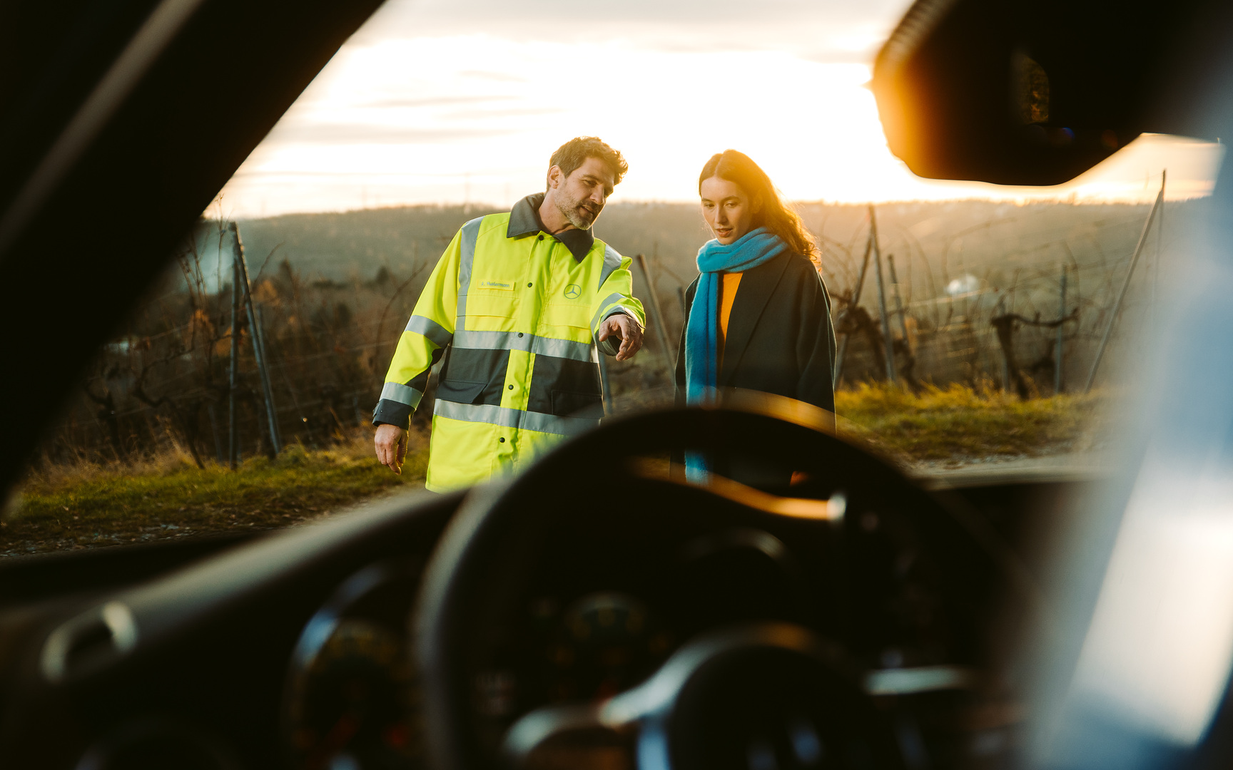 Assistance en cas de panne et de sinistre | Mercedes-Benz A travers le pare-brise d'une Mercedes, nous voyons un technicien de service qui se tient à côté de la conductrice devant le véhicule et le montre du doigt en guise d'explication.