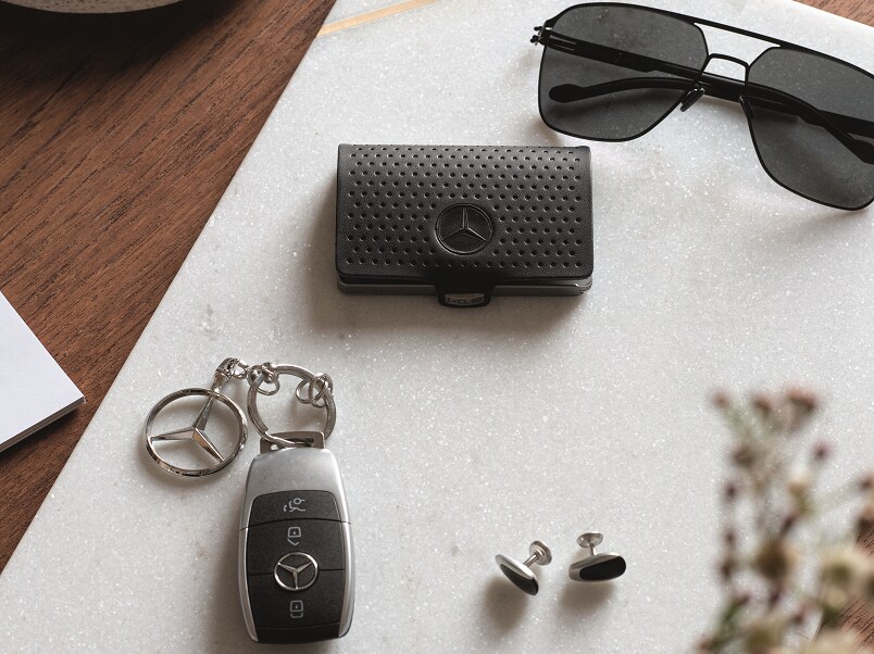 Une clé Mercedes avec un pendentif représentant l'étoile Mercedes-Benz, des boutons de manchette, des lunettes de soleil noires et un porte-monnaie se trouvent sur une tablette en marbre blanc posée sur une table en bois sombre.