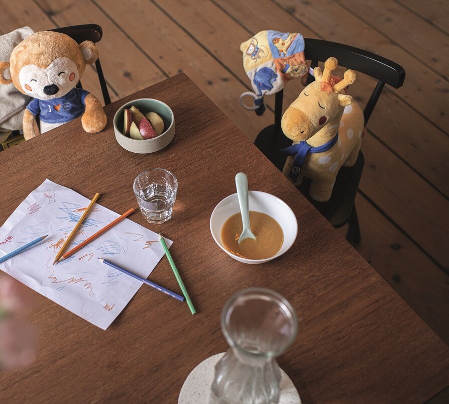 Des animaux en peluche sont assis sur des chaises autour d'une table sur laquelle se trouvent de la nourriture, de l'eau, des crayons et un dessin d'enfant commencé.