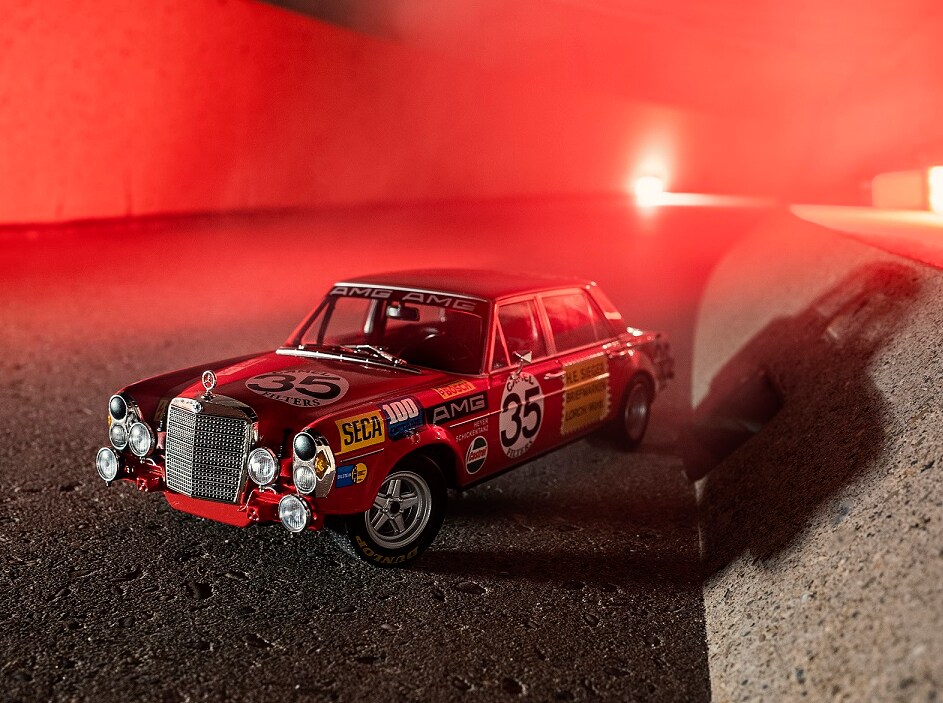 Une Mercedes 300 SEL AMG de 1971 avec une peinture authentique de voiture de course se tient en modèle réduit devant une lumière rouge éclatante.