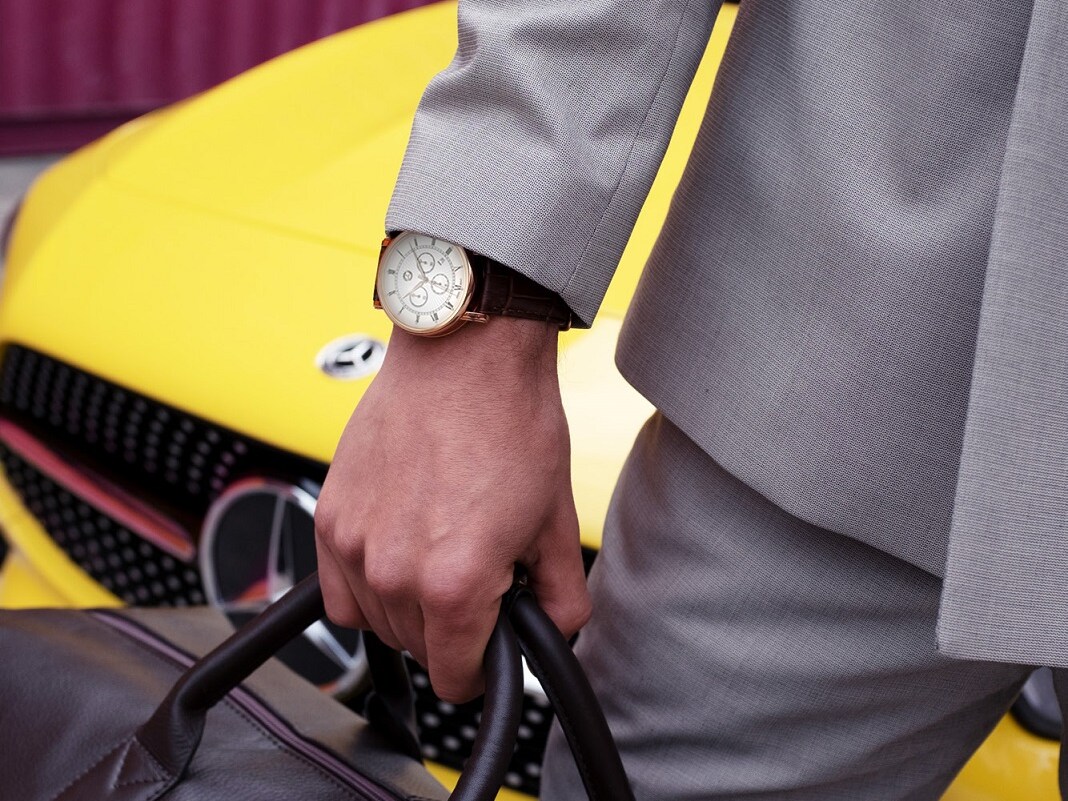 Cet extrait de photo montre un homme qui se tient devant une Mercedes jaune avec un sac de la Collection Mercedes-Benz et une montre Mercedes-Benz au poignet.