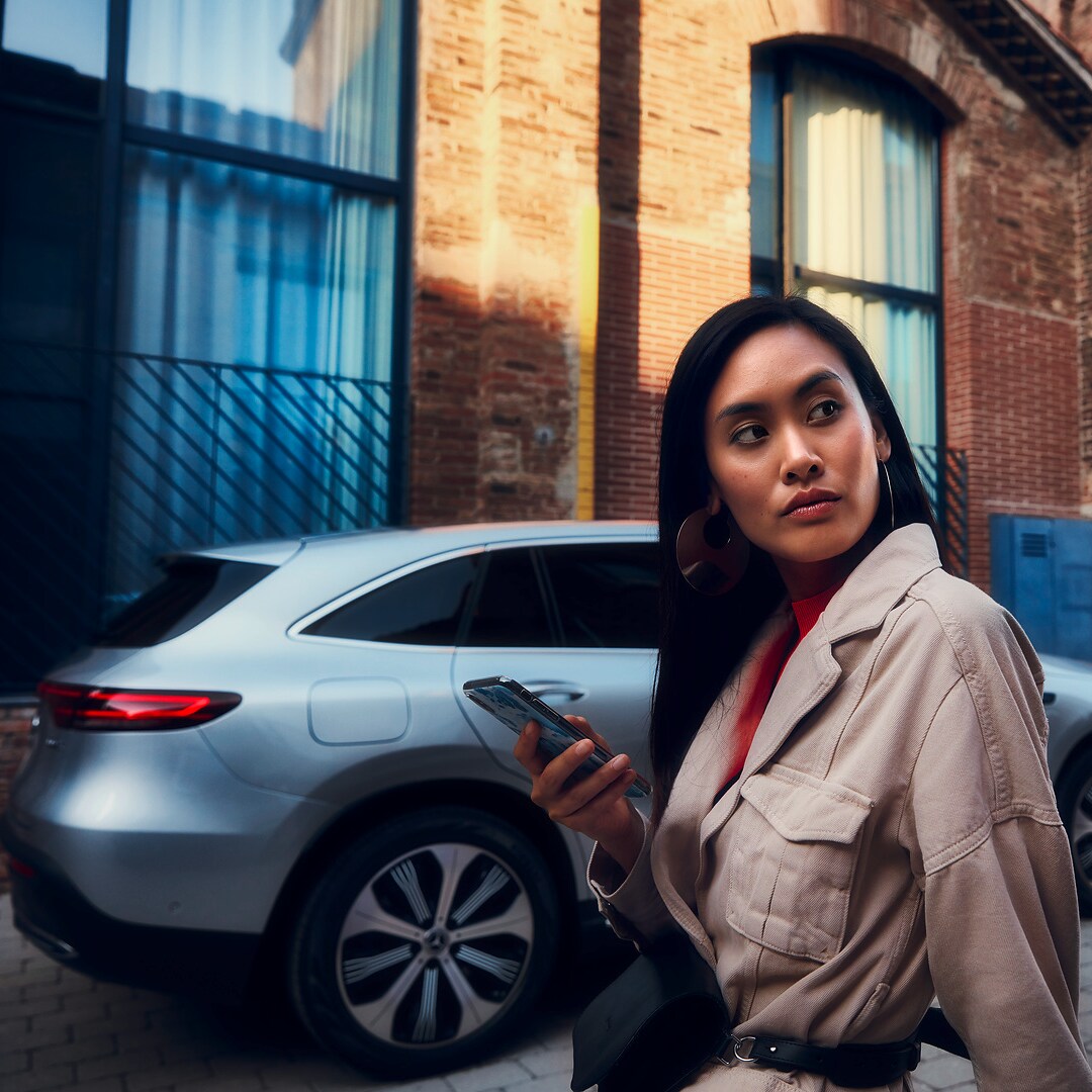 Une femme est debout devant sa Mercedes et tient un smartphone dans sa main droite.