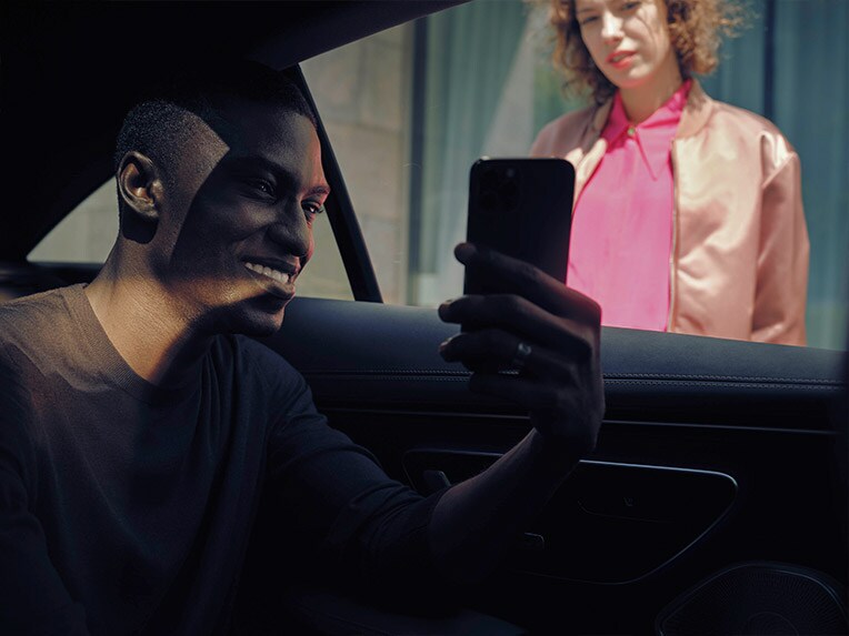 Un homme est assis dans une Mercedes-Benz et montre son téléphone portable à une femme qui se trouve à l'extérieur