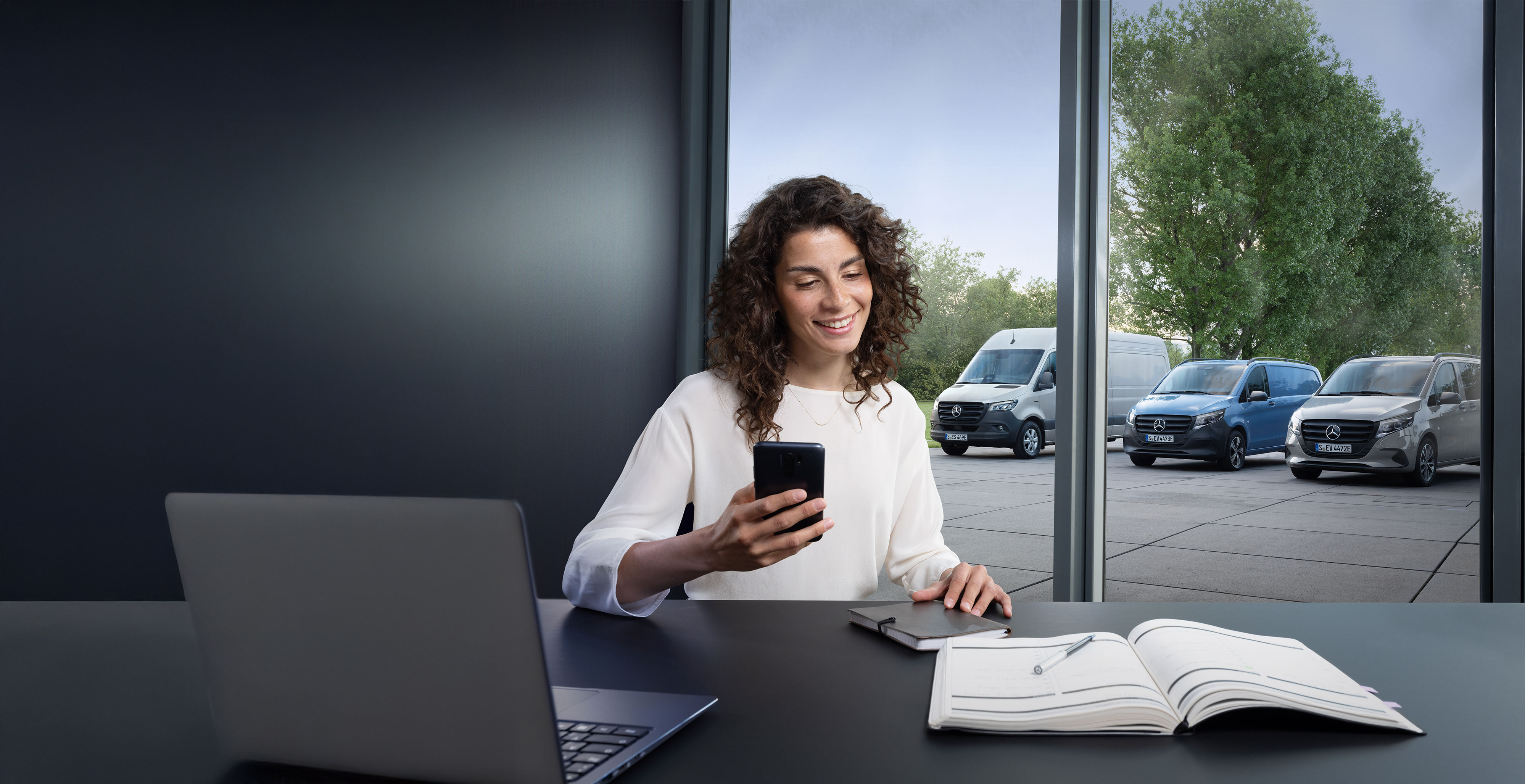 Mercedes-Benz | Maintenance des utilitaires légers Femme avec smartphone dans un bureau, plusieurs utilitaires légers Mercedes-Benz à l'arrière-plan - soutien d'experts dans le domaine de l'assistance aux entreprises.