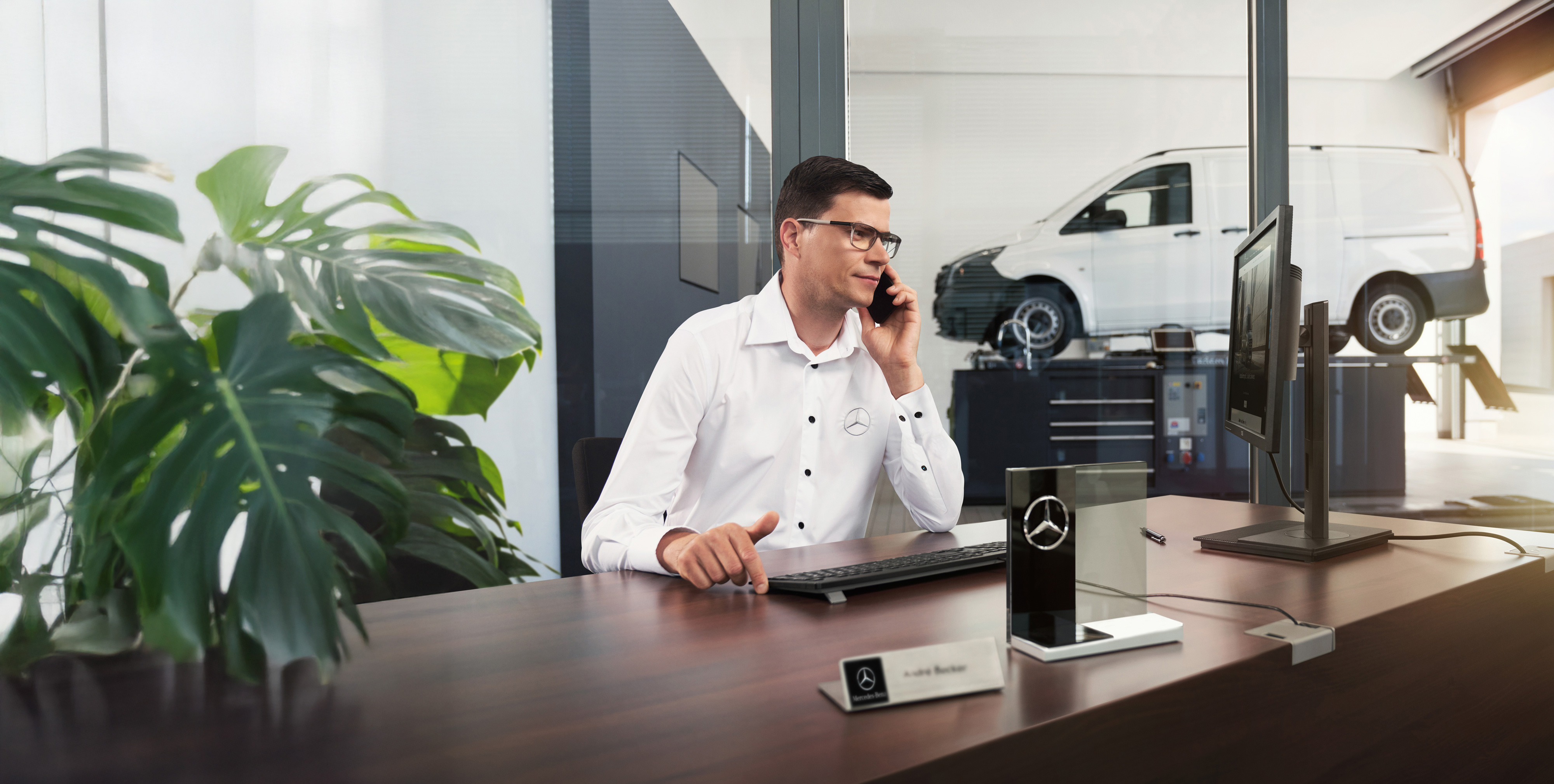 Mercedes-Benz | Maintenance des utilitaires légers Un employé de Mercedes-Benz parle au téléphone à un bureau, alors qu’un utilitaire léger Mercedes-Benz Vito se trouve dans l'atelier.