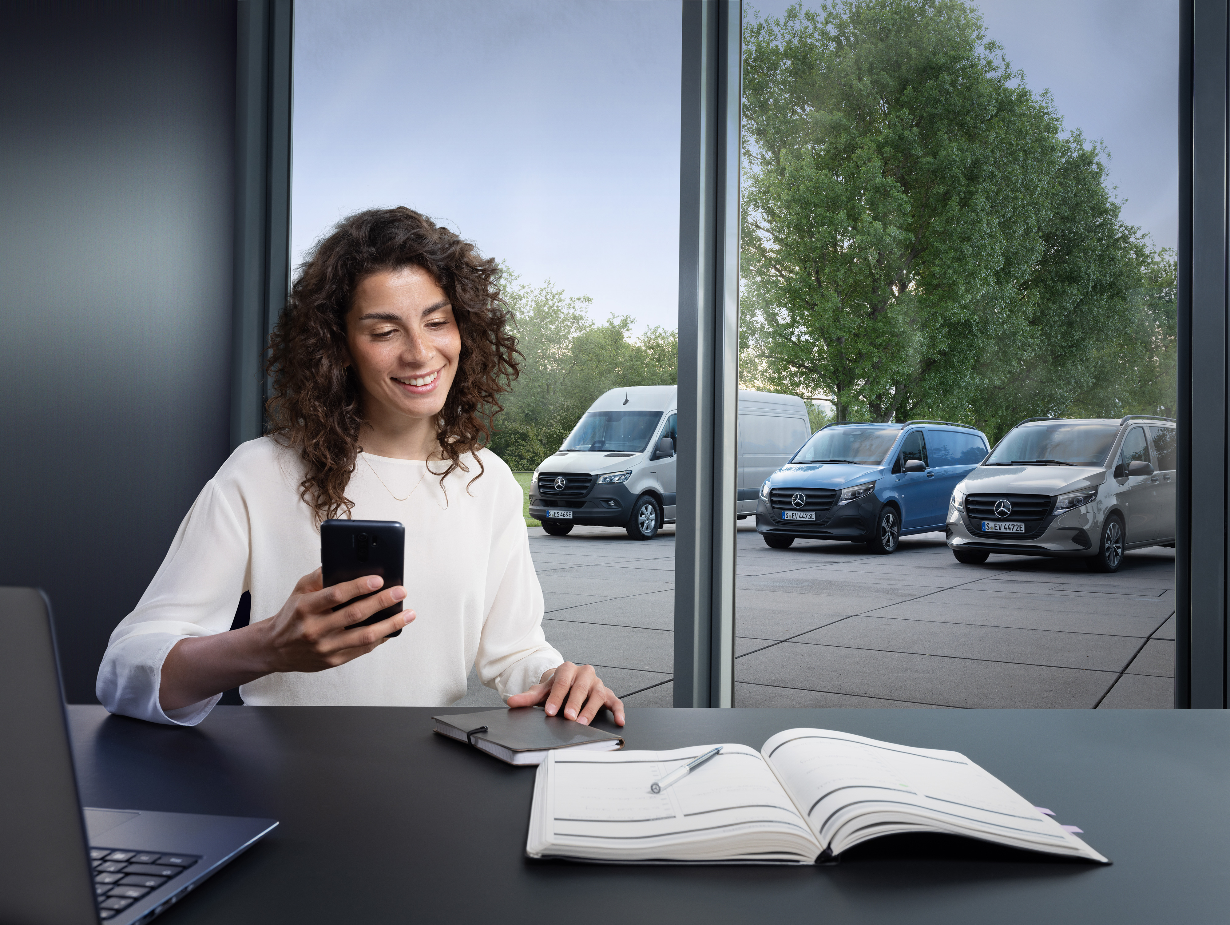 Mercedes-Benz | Van ServiceCare | Appointment booking Woman planning on her smartphone in front of a range of Mercedes-Benz vans in the background.